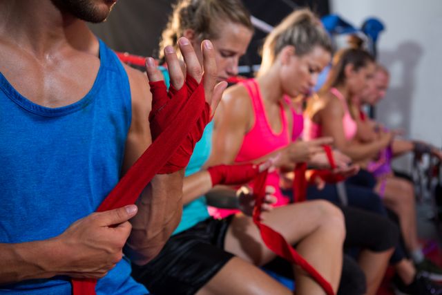 Group of athletes sitting in a row, wrapping red bandages around their hands in a fitness studio. Ideal for use in articles or advertisements related to fitness, boxing training, sports preparation, teamwork, and active lifestyles.