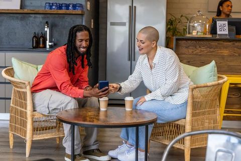 Friends sharing smartphone and coffee while relaxing in cozy cafe on wicker chairs