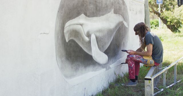 Street artist sitting and taking a break next to partially completed dolphin mural. They are holding a smartphone, wearing casual attire, and surrounded by painting supplies. Use this to depict urban creativity, modern street art, or promoting artistic events or communities.