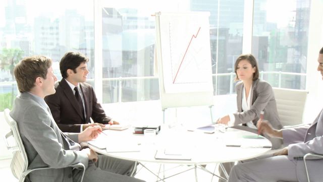 With a backdrop of a modern office setup, the image highlights diverse business professionals actively engaging in a team meeting or conference. They are analyzing graphs, implied by the flip chart in their midst. This setting fosters concepts of collaboration, brainstorming, and strategic planning effectively. Suitable for materials relating to office culture, teamwork presentations, or corporate interactions.