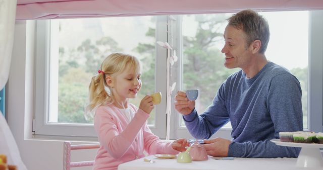 Father and young daughter enjoying tea party, smiling and bonding in front of large window with natural light. Great for themes of family, parenting, playtime, and nurturing relationships, ideal for advertisements, parenting blogs, and children's activity guides.