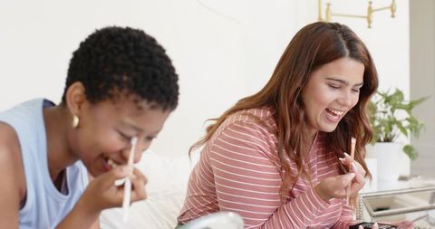 Friends applying makeup with brushes and palette laughing on bed cozy morning routine