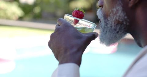 Senior african american man sipping cocktail by pool with strawberry, relaxed patio