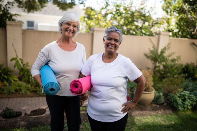 Two senior women are standing outdoors in a park, each holding a yoga mat and smiling. They are dressed in casual exercise clothing, suggesting they are ready for a fitness session. This image can be used for promoting senior fitness programs, healthy lifestyles, outdoor activities, and wellness among older adults.