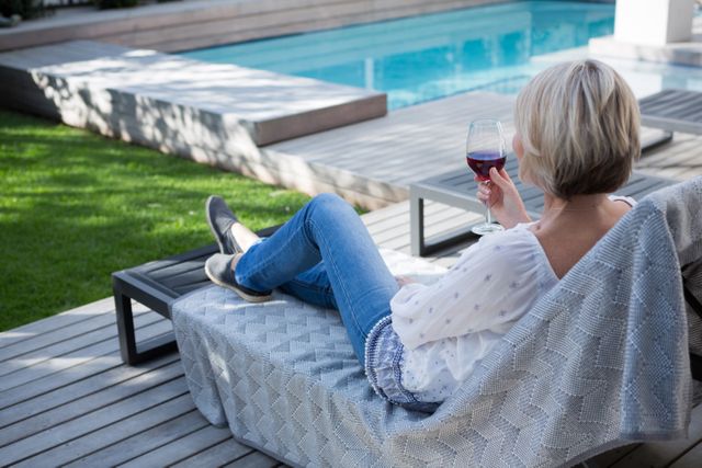Woman sitting on a lounge chair by the pool, enjoying a glass of wine on a sunny day. Ideal for use in lifestyle blogs, advertisements for outdoor furniture, wine promotions, or articles about relaxation and leisure activities.