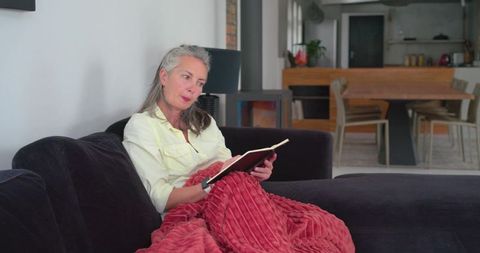 Contemplative woman reading notebook on sofa wrapped in red throw in modern home
