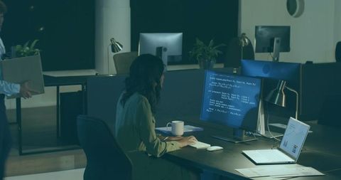 Female developer coding at night in modern office workspace with dual screens and charts