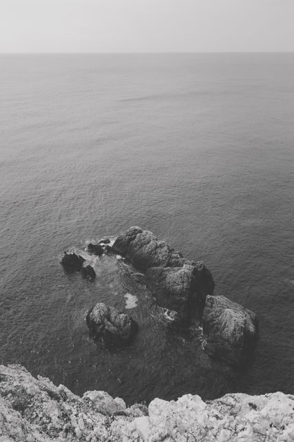 Coastal rocks can be seen in the vast sea with waves gently breaking against them in a black and white photograph. This image emphasizes the natural beauty of the ocean and the rugged landscape, creating a serene and tranquil atmosphere. Ideal for use in travel blogs, nature photography showcases, and tranquil motivators, it can also be used for wall art in minimalist designs or meditative spaces.