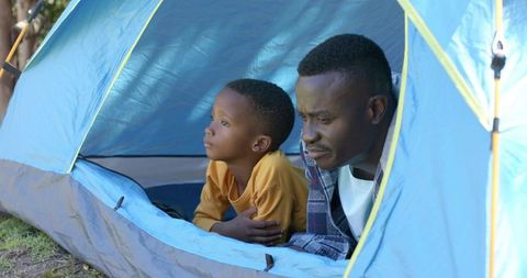 African american father and son camping in backyard tent peering out, family bonding