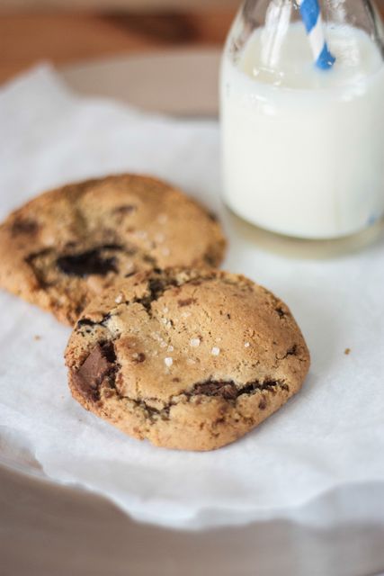Close-up view of two freshly baked chocolate chip cookies on parchment paper, accompanied by a bottle of milk with a blue straw. Perfect for illustrating homemade snacks, baking recipes, or comfort food in blogs, websites, and cookbooks. This image highlights concepts of indulgence, simplicity, and classic dessert enjoyment.