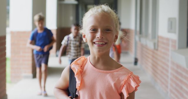 Young blonde girl smiling while walking in a school hallway alongside friends, carrying backpacks. Useful for educational content, back-to-school promotions, and children's lifestyle advertisements.