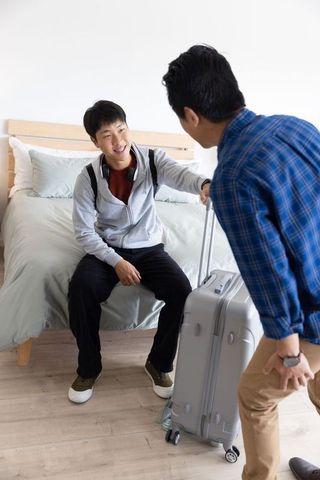 Asian traveler arriving and greeting roommate while pulling suitcase in bright bedroom