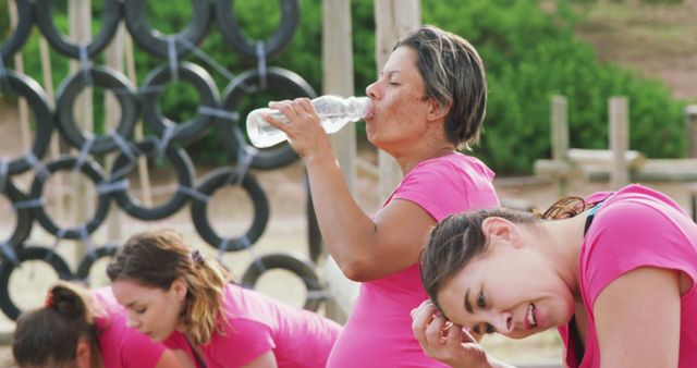 Tired caucasian female friends in pink t shirts resting and drinking water at bootcamp training. Female fitness, challenge and healthy lifestyle.