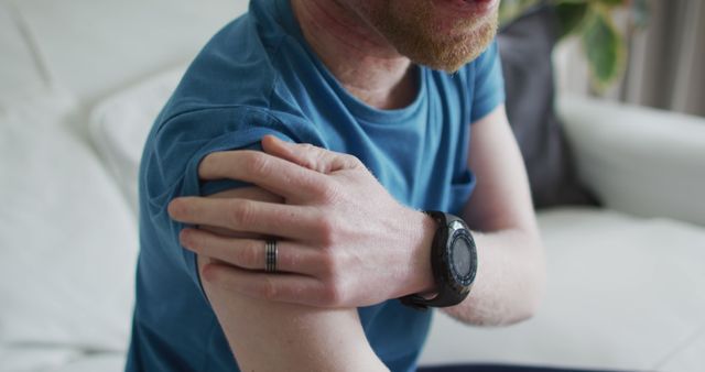 Man in casual blue shirt massaging his sore arm while sitting on a couch. He wears a smartwatch and a ring. This image can be used for content related to muscle pain relief, healthcare advice, relaxation techniques, or smartwatch functionality.