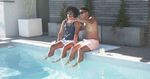 Young men relaxing by pool dipping feet and pointing while sunbathing on modern patio