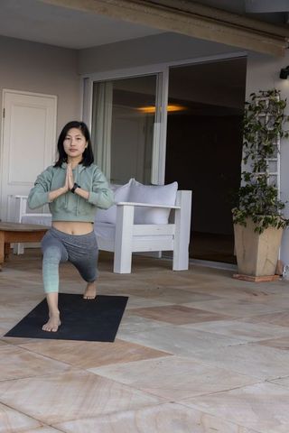 Asian woman practicing yoga lunge on tiled patio wearing smartwatch, stretching, meditating