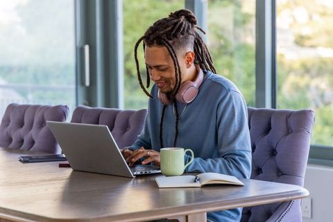 African american man working on laptop at home, wearing pink headphones with coffee