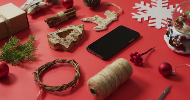 This festive holiday image showcases a desktop with various Christmas decorations including a gift, wooden ornaments, pine branches, a snowflake decoration, cinnamon sticks, ribbon, a smartphone, and crafting materials on a red background, ideal for Christmas promotions, holiday blogs, or social media posts.