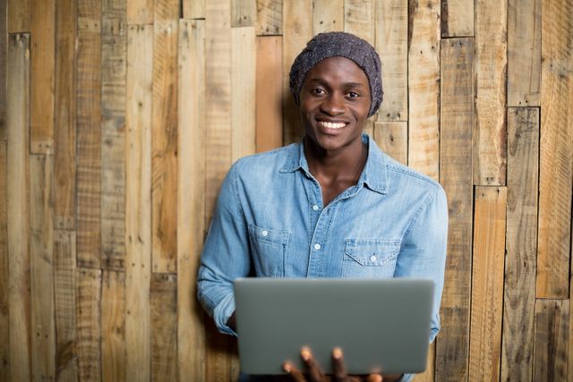 Young African American man in denim shirt and beanie smiling while using laptop against wooden wall. Ideal for illustrating concepts of remote work, freelancing, modern lifestyle, and technology use in casual settings.