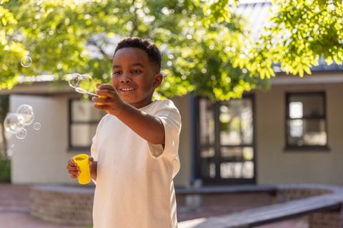 African american boy blowing soap bubbles in sunlit courtyard, joyful outdoor play