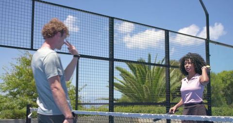 Casual players exchanging serve on sunny outdoor court with fence and lush greenery