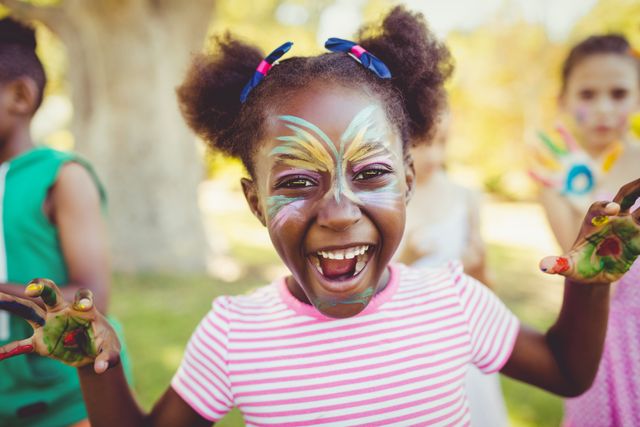 Girl with vibrant face paint and painted hands enjoying a fun day in the park. Perfect for themes related to childhood, outdoor activities, creativity, and joyful moments. Ideal for use in advertisements, educational materials, and family-oriented content.