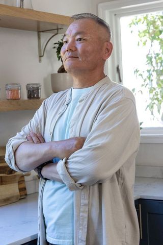 Mature asian man standing with arms crossed in bright modern kitchen, contemplative portrait