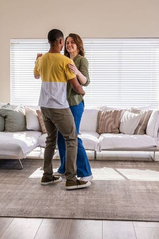 Sunlit diverse couple dancing together in cozy modern living room embracing