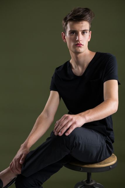 This image features an androgynous man sitting on a stool against a green background. He is wearing a black shirt and dark pants, exuding a casual yet stylish vibe. This photo can be used for fashion editorials, modern lifestyle blogs, or advertisements focusing on contemporary and gender-neutral fashion.