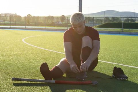 Male field hockey player tying shoe on turf pitch at sunset