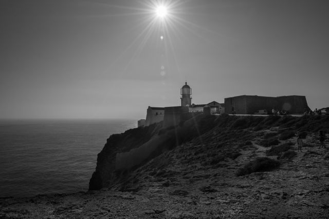 Black and white coastal landscape showing a sunlit lighthouse standing on a rocky cliff edge overlooking the ocean. Ideal for themes related to architecture, nature, coastal scenery, guidance, and nautical subjects.