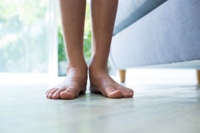 This image shows a child's legs and feet standing barefoot on a floor next to a sofa in a home setting. The bright and airy environment suggests a comfortable and relaxed atmosphere. This image can be used for themes related to childhood, home life, comfort, and casual living. It is ideal for use in advertisements, blogs, and articles focusing on family, home decor, and lifestyle.