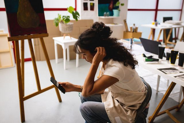 Biracial female artist with painting equipment and smartphone in creative office. Female artist studio and painting concept.