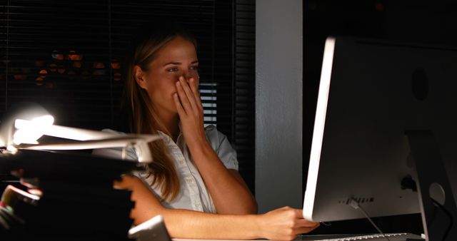Young woman is working late at an office desk, illuminated by a desk lamp and computer screen. She appears focused and tired as she continues her work with paperwork and a computer. This image illustrates the dedication and commitment of professionals, suitable for use in articles about work-life balance, productivity tips, or business commitment.