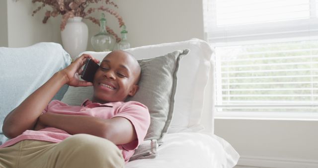 Young boy is comfortably lying on a couch and happily talking on his phone. Perfect for depicting scenes of modern communication, relaxed home environments, and child leisure activities. Can be used in advertisements for telecommunications, lifestyle magazines, or family-oriented content.
