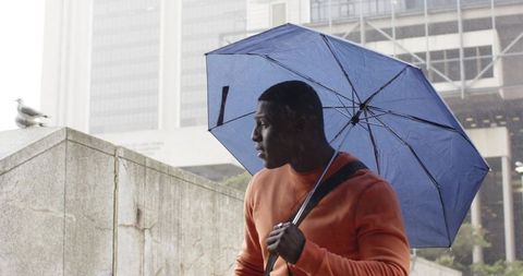 African American Man Holding Blue Umbrella Walking on Urban Ramp in Rain Wearing Orange Sweater