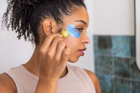 African woman using gold facial roller over blue under-eye patches during skincare routine