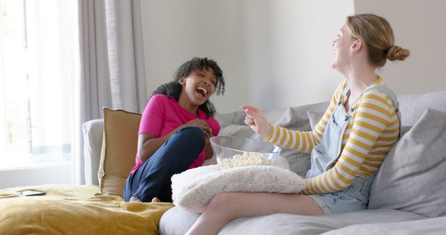 Two women are relaxing on a sofa, engaging in a cheerful conversation and sharing a bowl of popcorn. This setting signifies friendship, leisure time, and relaxation at home. Ideal for use in lifestyle blogs, social media posts about friendships, or advertisements promoting leisure and comfort at home.