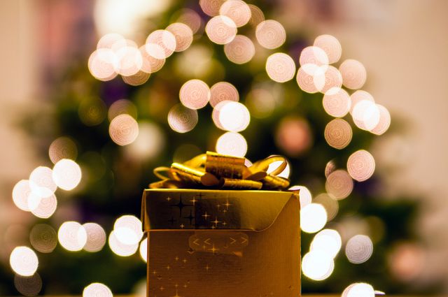 Golden gift box with a ribbon is positioned prominently against blurred bokeh Christmas lights. The holiday season theme shines through with festive decorations and the soft, glowing background. Ideal for holiday greeting cards, celebratory advertisements, festive content, and Christmas promotions.