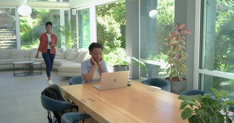 Teen working on laptop at modern sunlit dining table while caregiver walking by