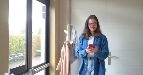 Woman standing by entryway checking smartphone while wearing glasses and denim shirt