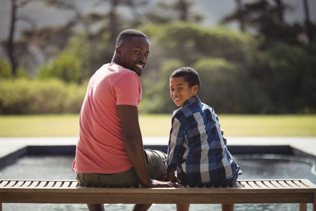 Father and son enjoying quality time together outdoors, sitting on a bench and smiling at the camera. Ideal for use in family-oriented advertisements, parenting blogs, and articles about father-son relationships. Suitable for promoting outdoor activities, family bonding, and leisure time.