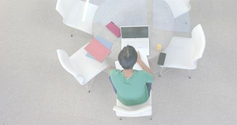 Overhead Asian student studying on laptop at round glass table campus lounge