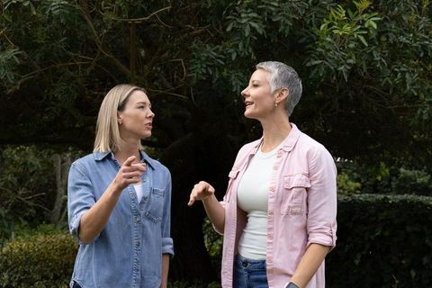 Two women chatting and pointing under garden tree, casual outdoor conversation