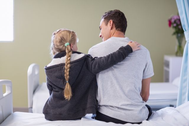 This image depicts a touching moment of a daughter comforting her sick father in a hospital room. It can be used in healthcare and family-related content, emphasizing themes of support, love, and recovery. Ideal for articles, blogs, and promotional materials focusing on family care, patient recovery, and emotional support in medical settings.