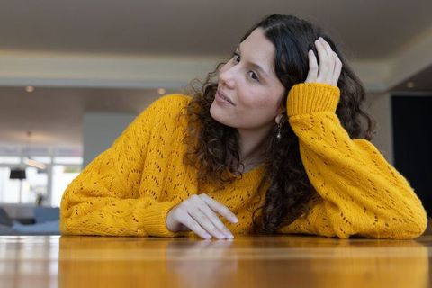 Young woman leaning on wood table in mustard sweater touching hair in bright modern lounge