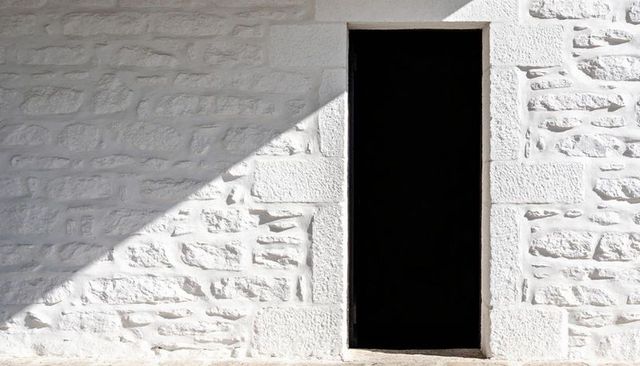 Narrow doorway framing black void in sunlit white stone masonry facade with diagonal shadow