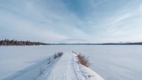 Drone flying over narrow snow trail crossing frozen lake toward distant mountain range