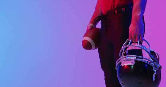 Football player standing with football helmet in one hand and holding football in the other, illuminated by vibrant purple, blue, and red neon lights. Perfect for sports articles, athlete portraits, training programs, and football promotional materials.