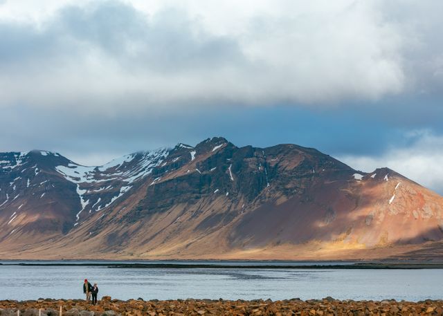 Two hikers walking along rocky shore with snow-capped mountain range and lake in background under cloudy sky. Ideal for use in travel blogs, adventure magazines, promotional tourism material, environmental campaigns.